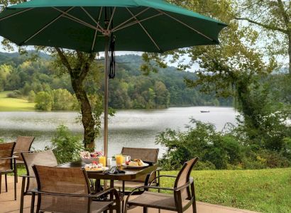 Outdoor dining setup with a table, chairs, umbrella, and breakfast spread by a lake with trees and hills in the background.