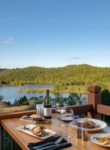 A scenic view overlooks a lush landscape and a lake. A table set with dishes, wine, and glasses offers a cozy dining spot on a balcony.