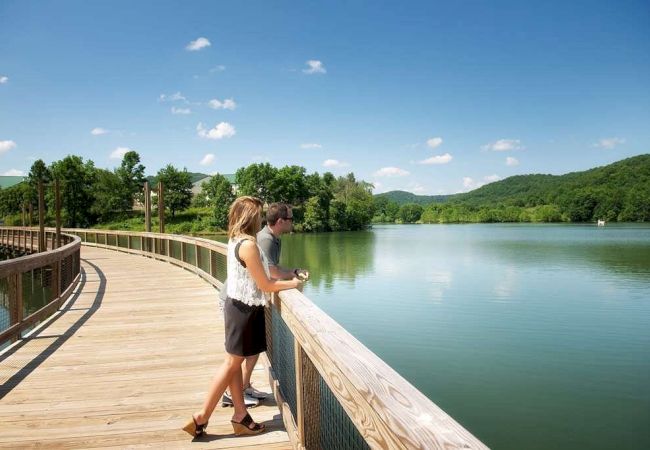 A couple stands on a wooden boardwalk overlooking a scenic lake surrounded by lush green hills and a clear blue sky.