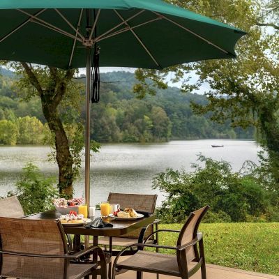 Outdoor dining table with food under a green umbrella, overlooking a scenic lake and trees.