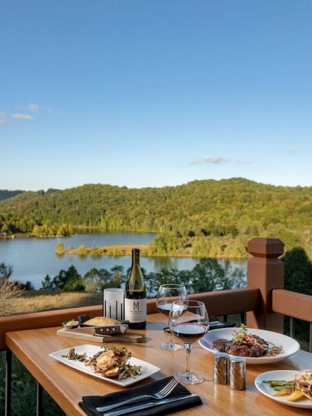 A table with plates of food and wine overlooks a scenic view of a lake and lush, green hills under a clear blue sky.
