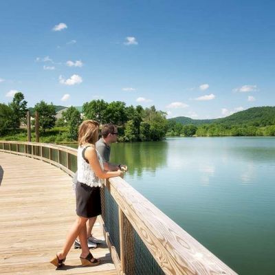 A couple stands on a wooden boardwalk overlooking a calm lake, surrounded by lush green hills and a clear blue sky with scattered clouds.