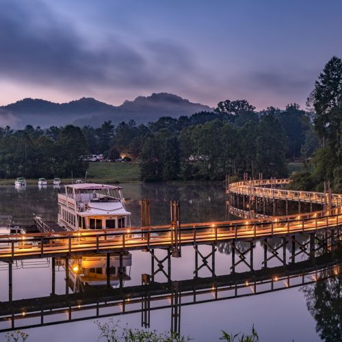 A serene view of a boat on a lake, surrounded by trees with a lit-up boardwalk, and mountains in the background at dusk.