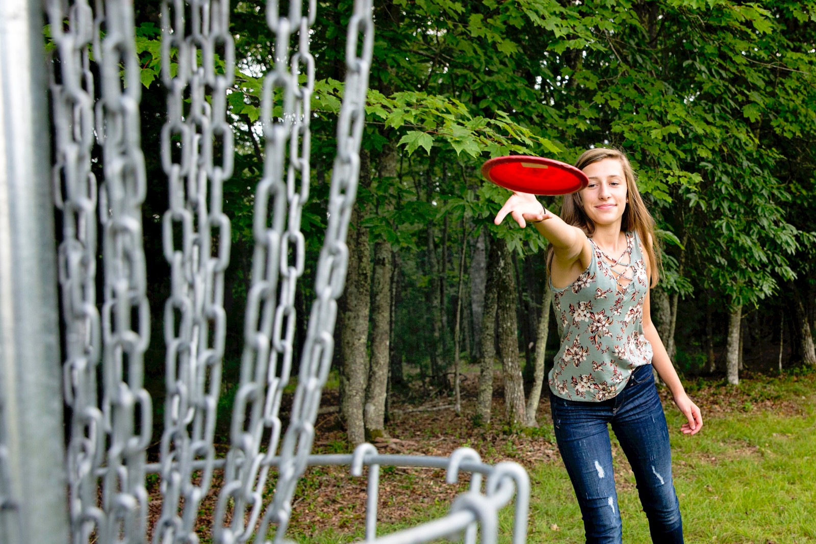 A person playing disc golf, aiming a red disc toward a metal basket, surrounded by trees in the background.