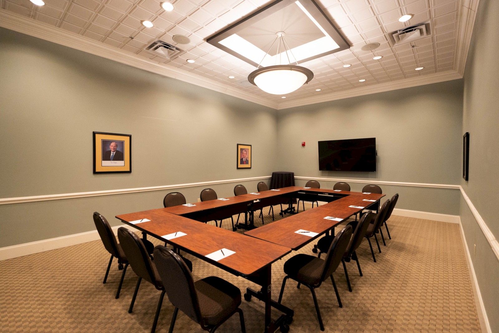 A conference room with a U-shaped table layout, chairs, a TV, and framed photos on mint green walls, well-lit by ceiling lights.
