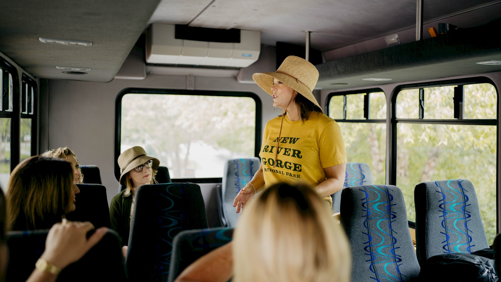 A woman wearing a yellow shirt and hat is speaking to a small group of seated people inside a bus.