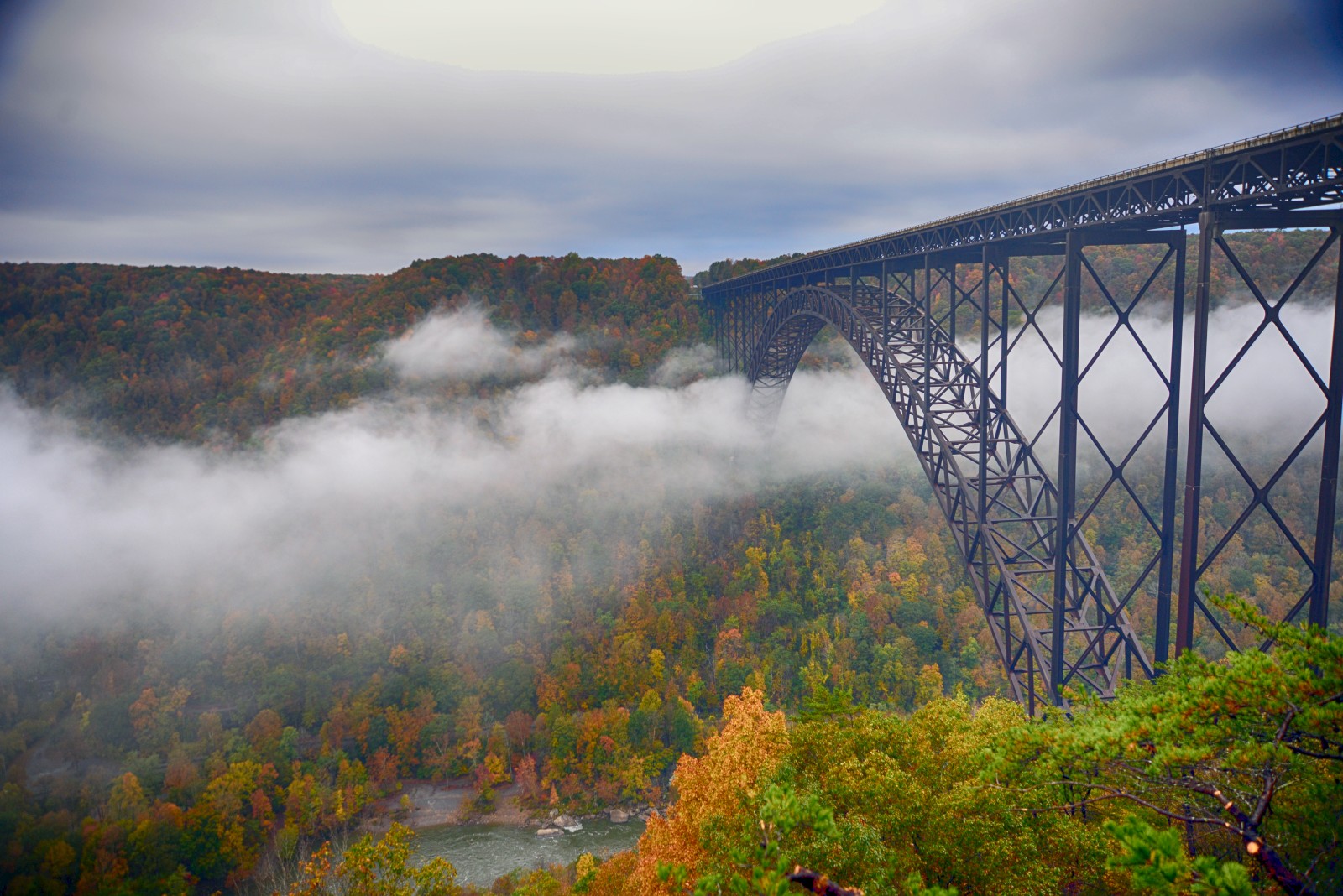 A tall steel arch bridge spans a misty river gorge, with autumn trees in orange and yellow below. The scene is moody and scenic, suspended in fog.