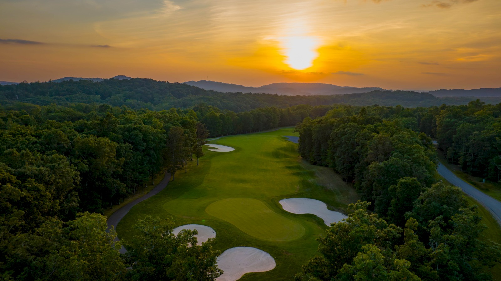 A scenic golf course at sunset, with lush greenery, sand traps, and a winding path surrounded by trees and hills in the distance.