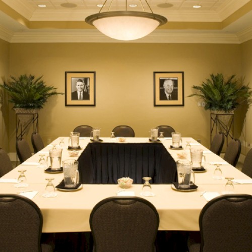A conference room with a U-shaped table arrangement, framed photos on the walls, plants in corners, and chairs set for a meeting.