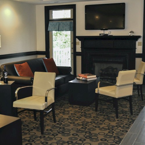 A cozy living room with a fireplace, TV, sofas, and armchairs, featuring a modern design and patterned carpet, lit by a standing lamp.