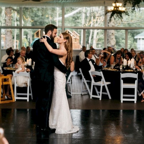 A couple is dancing at a wedding reception, surrounded by seated guests in an elegantly decorated venue.