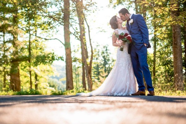 A bride and groom share a tender moment on a sunlit path surrounded by trees, with the bride holding a bouquet of red and white flowers.