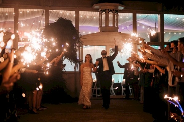 A couple in formal attire walks through a crowd holding sparklers, celebrating at night outside a lit-up venue.