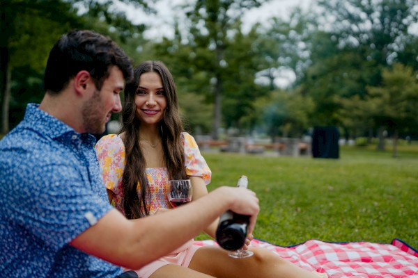 A couple having a picnic on a red-checkered blanket, enjoying wine, surrounded by trees in a park setting.