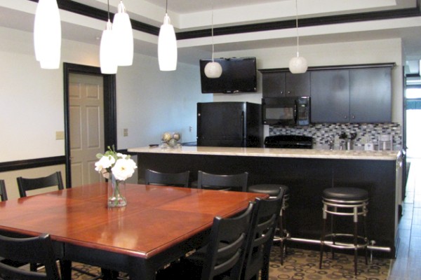 This image shows a modern kitchen and dining area with black cabinets, a wooden table, bar stools, and pendant lights.