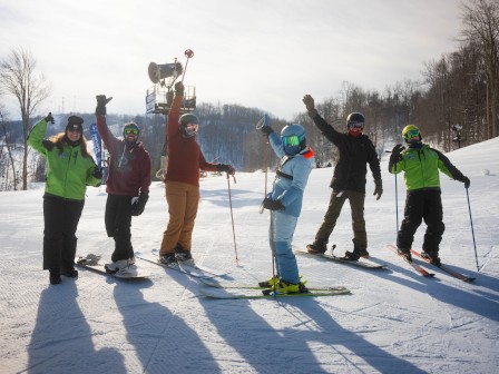 A group of friends on a sunny snowy slope celebrate a win with raised arms and a trophy, all wearing colorful ski gear and poles.