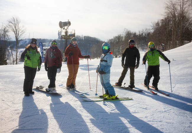 A group of six people in ski gear standing on a snowy slope, with ski poles, under a clear sky and surrounded by trees.