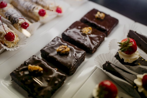 The image shows a variety of desserts, including chocolate brownies with nuts, and pastries topped with cherries and strawberries on a white tray.