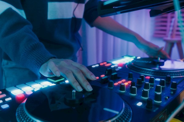A DJ is mixing music, using a turntable with lit buttons and controls, creating an energetic atmosphere in a dimly lit room.