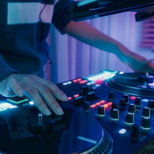 A person is DJing, using a turntable with lit-up buttons and knobs in a dimly lit setting.