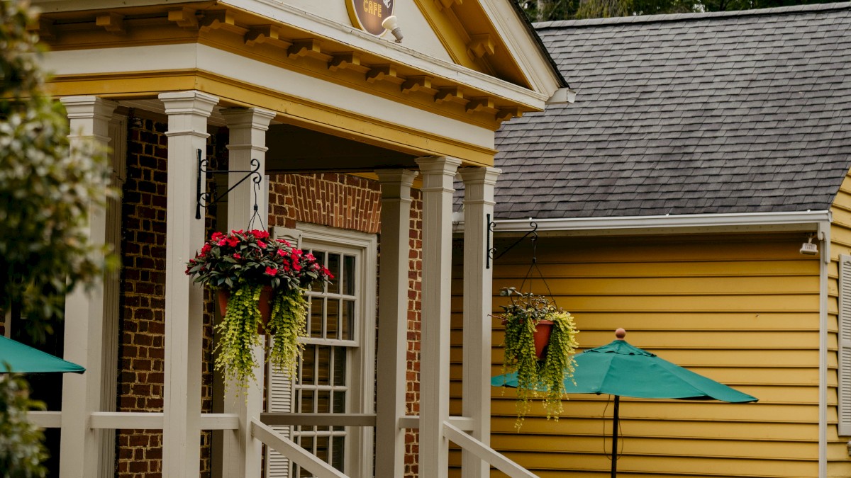 A charming yellow house with a wooden porch, hanging flowers, and umbrellas outside, surrounded by greenery and a cobblestone path.