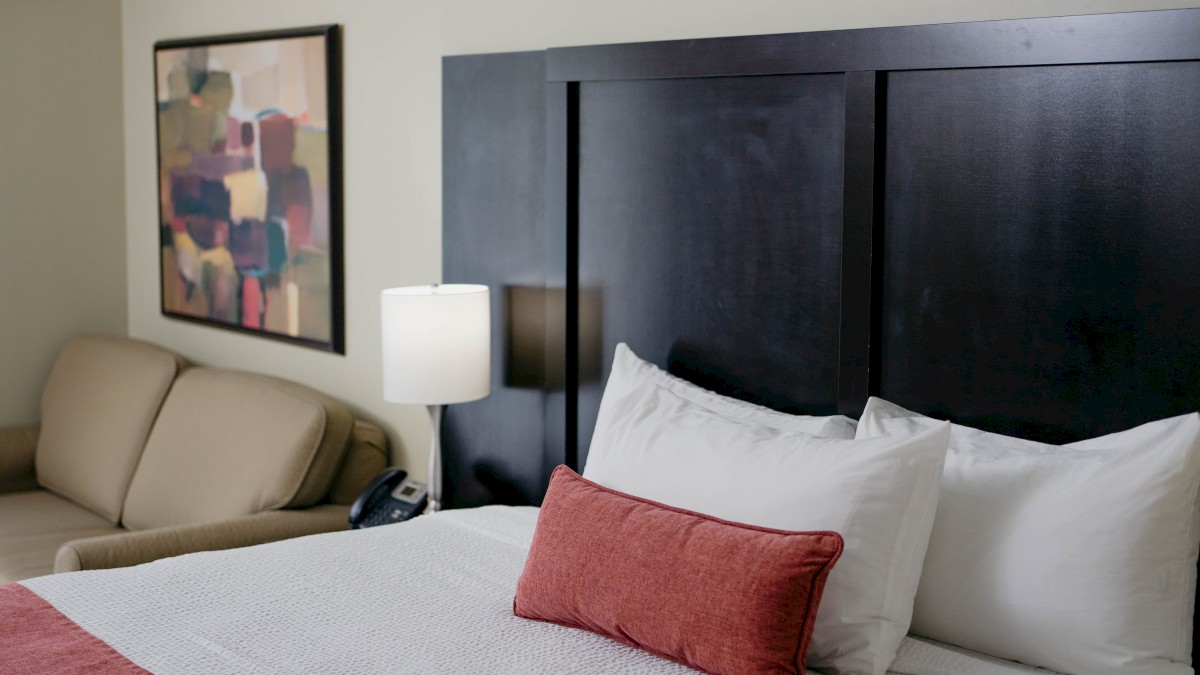 A hotel room features a bed with red accents, a sofa, a lamp, and abstract art on the wall.