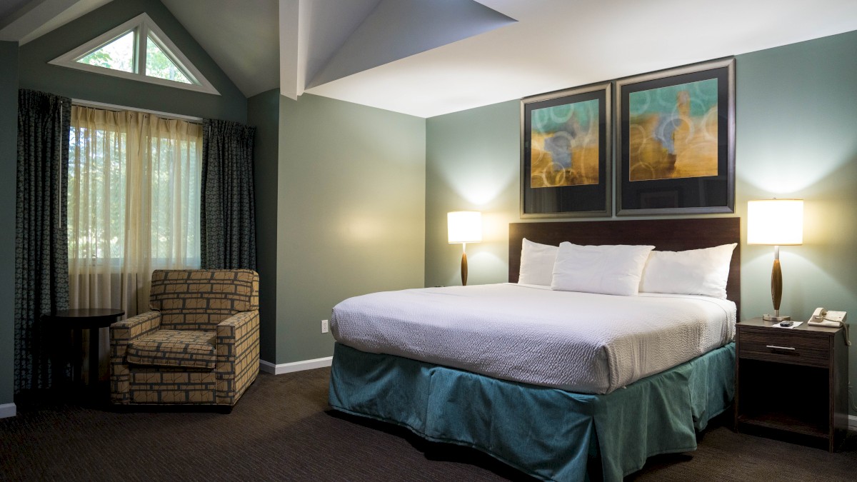 The image shows a hotel room with a king-sized bed, two bedside lamps, artwork above the bed, an armchair, and large window curtains.