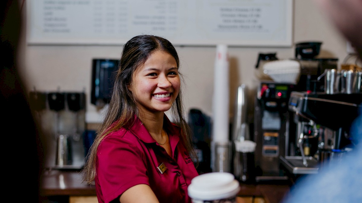 A barista in a red shirt hands a cup of coffee to a customer at a café counter with a menu board in the background.