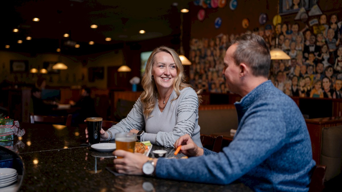 Two people sitting at a bar, enjoying drinks and food while talking, with a colorful wall mural in the background and warm lighting.