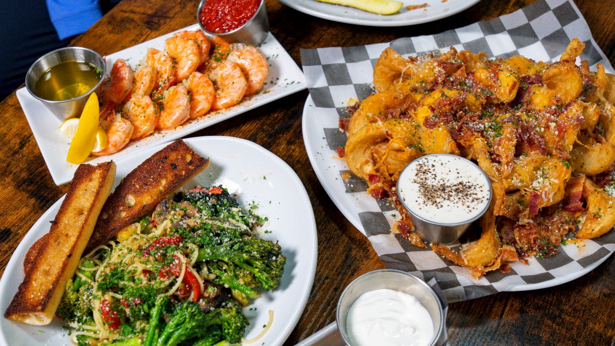 The image shows a wooden table with several plates of food: shrimp, salad, cheesy bread, a burger, and potato wedges with sauces.