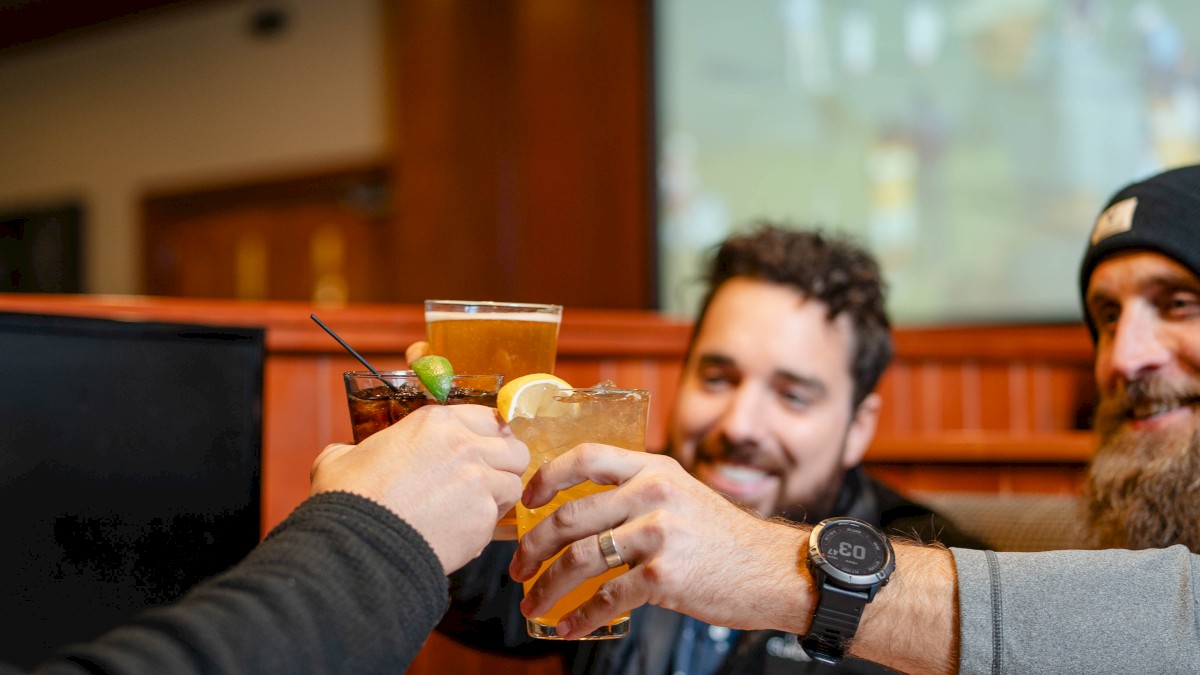 A group of people is clinking glasses at a restaurant table, with a screen visible in the background.