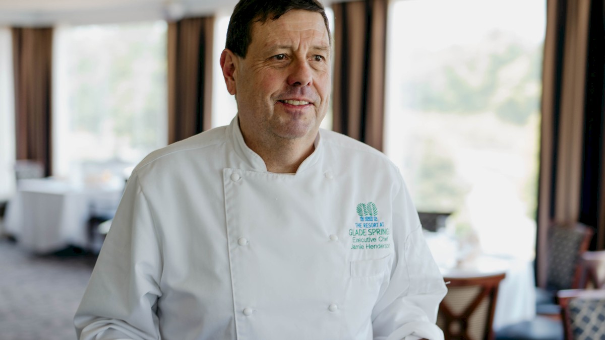 A chef in a restaurant is carrying two plates of elegantly presented food, standing in a dining area with large windows.
