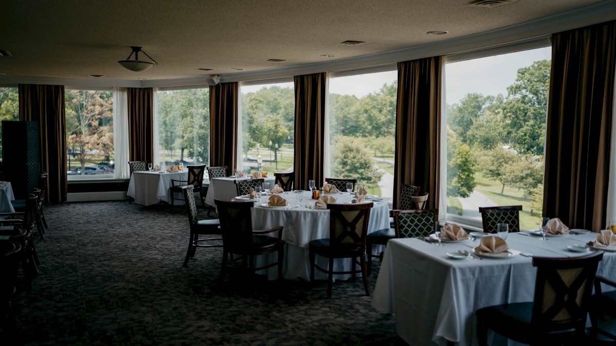 The image shows an elegant dining room with several round tables set for a meal, large windows offering a view of trees outside.