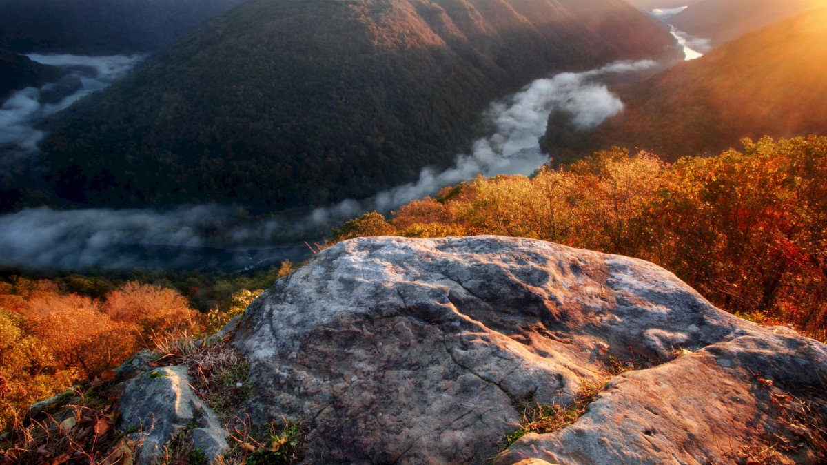 A scenic view of a rocky overlook with lush hills, misty river, and sunrise in the background, creating a serene and picturesque landscape.