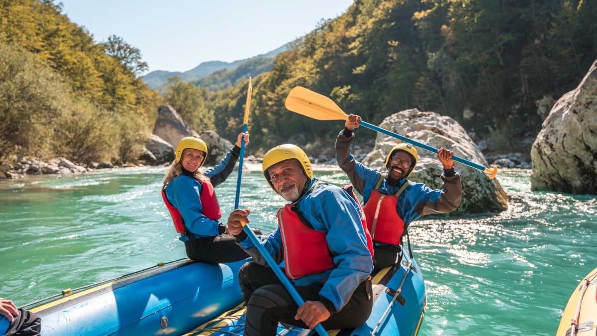 Three people in helmets and life jackets are joyfully rafting down a river, with trees and rocks in the background.