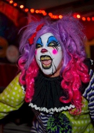 A person dressed as a scary clown with bright makeup, purple and pink hair, and a colorful costume in a dimly lit setting.