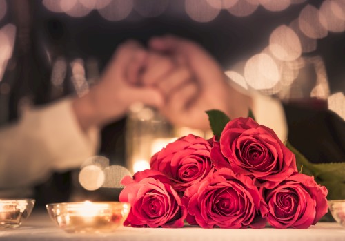 Romantic scene with a bouquet of bright pink roses on a table, blurred candlelight and a couple holding hands in the background.