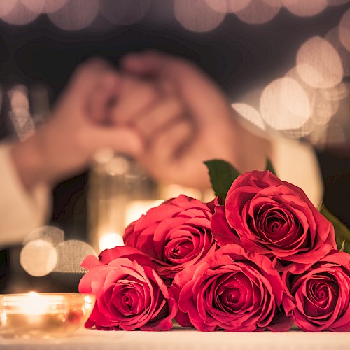 Romantic scene with a bouquet of bright pink roses on a table, blurred candlelight and a couple holding hands in the background.