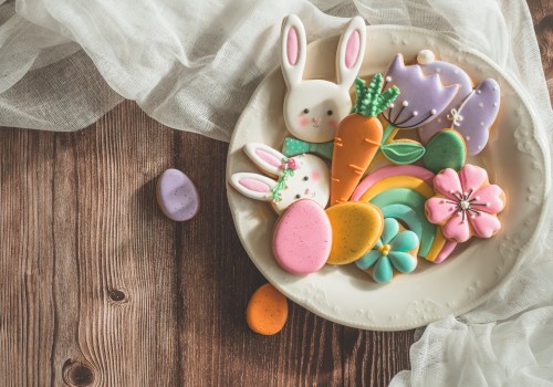 A plate with colorful Easter-themed cookies, including bunnies, carrots, flowers, and eggs, on a wooden table with fabric draped nearby.