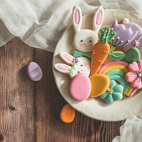 A plate with colorful Easter-themed cookies, including bunnies, carrots, flowers, and eggs, on a wooden table with fabric draped nearby.