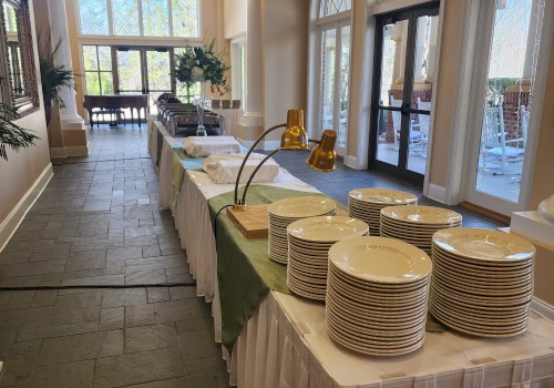A buffet setup with stacks of plates, long tables, and decorations in a well-lit room with large windows and arched features.