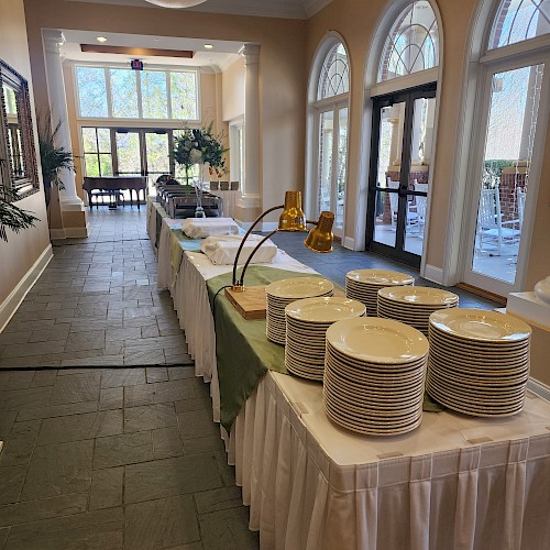 A buffet setup with stacks of plates, long tables, and decorations in a well-lit room with large windows and arched features.