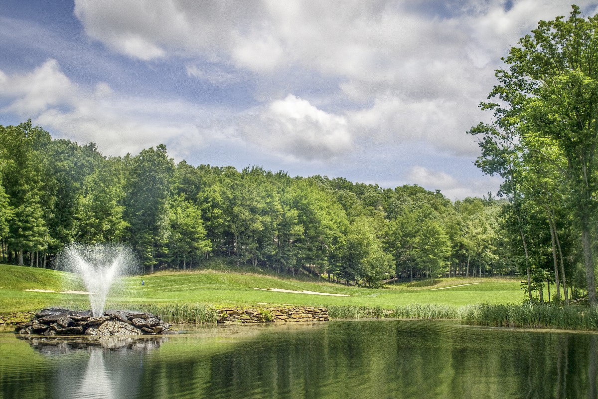 A tranquil park scene with a pond, a small fountain spray, lush green fairway, and a background of trees under a partly cloudy sky.