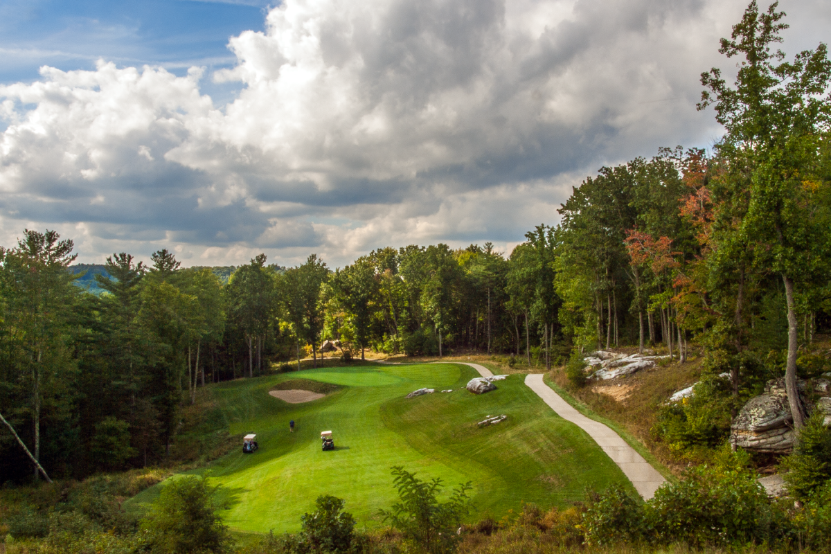 A scenic golf course tucked in a wooded valley, with a winding path, rolling greens, carts, and rocky outcrops under a partly cloudy sky.