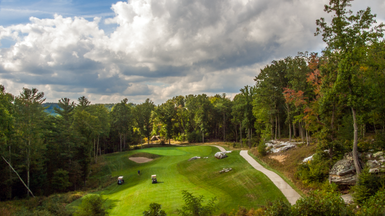 A scenic golf course tucked in a wooded valley, with a winding path, rolling greens, carts, and rocky outcrops under a partly cloudy sky.