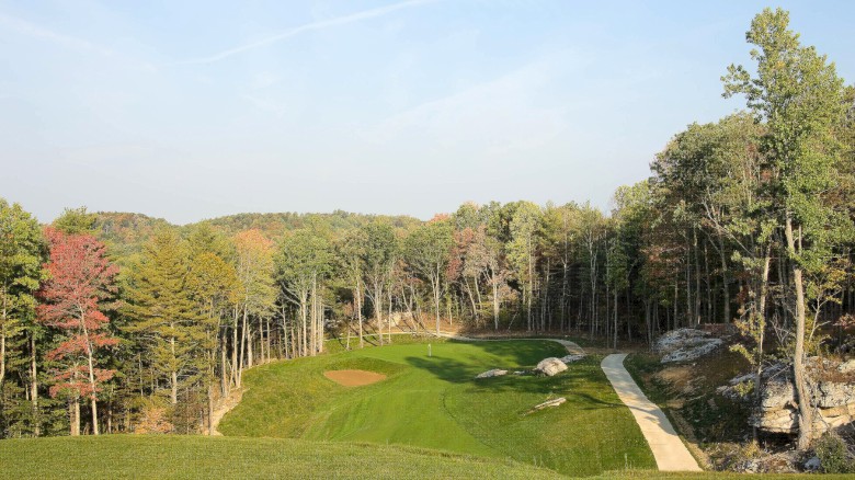 A scenic golf course set among trees with a green, fairway, bunkers, and a stone path on the right, under a clear blue sky.