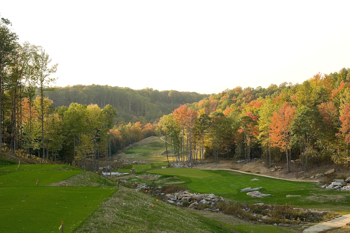A scenic autumn golf course winding through a forested valley with colorful fall foliage and rolling greens, under a bright sky.