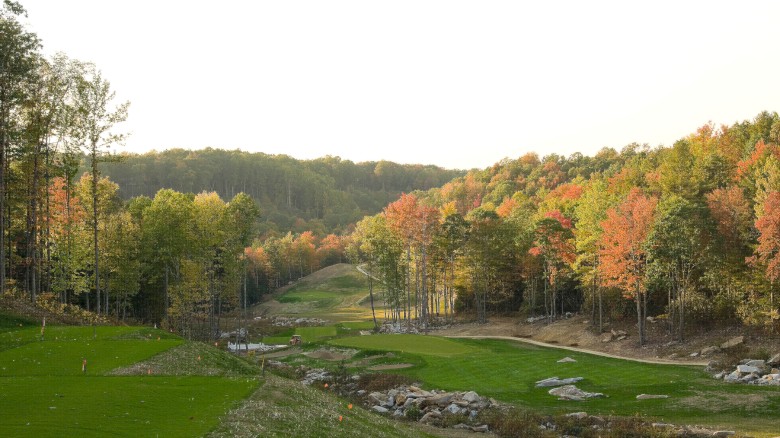 A scenic autumn golf course winding through a forested valley with colorful fall foliage and rolling greens, under a bright sky.