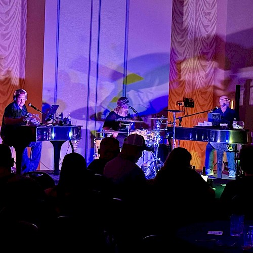 A band performs in a dimly lit room with an audience, featuring two keyboard players and a drummer on stage.