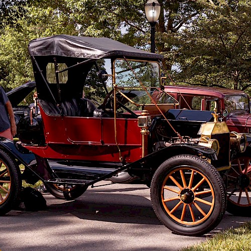 The image shows a vintage car with a red body and large wooden wheels on display outdoors, surrounded by trees and people.